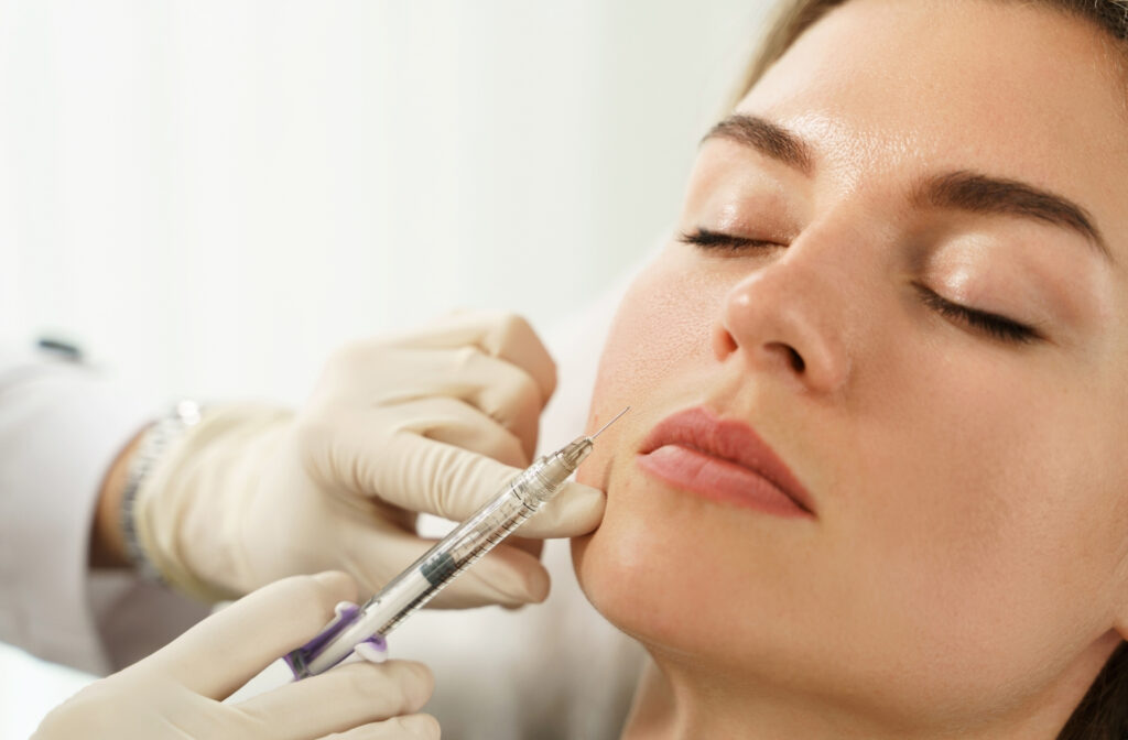 A close-up image showing a woman with her eyes closed receiving a cosmetic in her cheek. The practitioner is wearing white medical gloves and gently holds the woman's face while inserting a syringe with a fine needle into the cheek area. The setting is bright and clinical, highlighting a precise and careful cheek augmentation procedure.