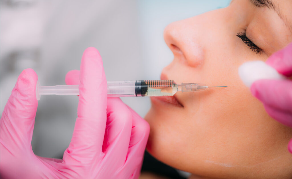 A close-up photo of a woman receiving a cosmetic injection in her cheek. A practitioner wearing bright pink gloves is holding a syringe filled with a clear substance, carefully injecting it into the woman's skin near her cheekbone. The woman's eyes are close, and a cotton pad is held near the injection site. The setting suggests a clinical or medical aesthetics environment focused on natural facial enhancement.